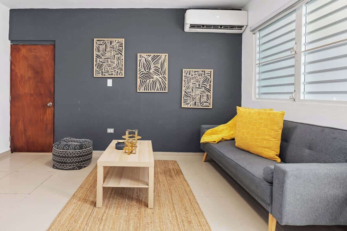 A living room with a dark wall, a wooden door, a gray sofa with a yellow pillow, a wooden coffee table, and three wall-mounted artworks.