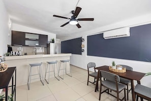 A modern kitchen with a central island, bar stools, a dining table with chairs, a ceiling fan, and an air conditioning unit.