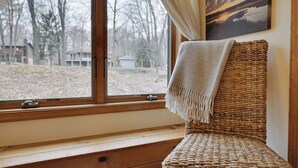 A wicker chair with a throw blanket, positioned by a window with a view of a wooded area. - Wooded Lakeside adventure, walk to dock, 4 kid kayaks, screened porch, ping pong (Pocono Lake)