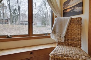 A wicker chair with a throw blanket, positioned by a window with a view of a wooded area. - Wooded Lakeside adventure, walk to dock, 4 kid kayaks, screened porch, ping pong (Pocono Lake)