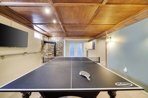 A ping pong table with a paddle in the center, a stone wall, and a flat-screen TV mounted on the wall. - Wooded Lakeside adventure, walk to dock, 4 kid kayaks, screened porch, ping pong (Pocono Lake)