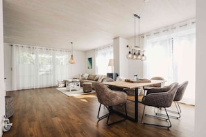 A spacious living room with a wooden floor, a sofa, a dining table with chairs, and a hanging light fixture.