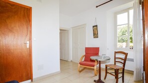 Quarto com porta de madeira, uma cadeira vermelha, uma mesa redonda e janela com vista para vegetação.