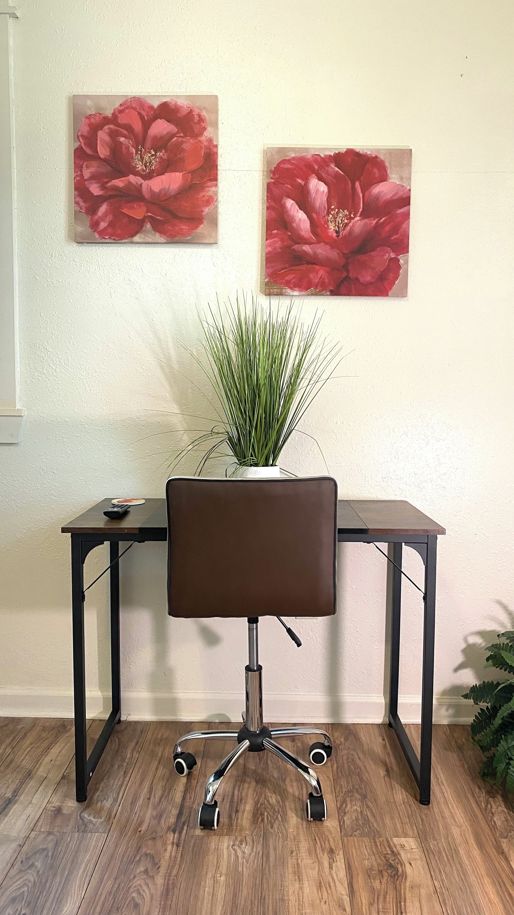 A desk with a brown chair, a potted plant, and two red flower paintings on the wall.