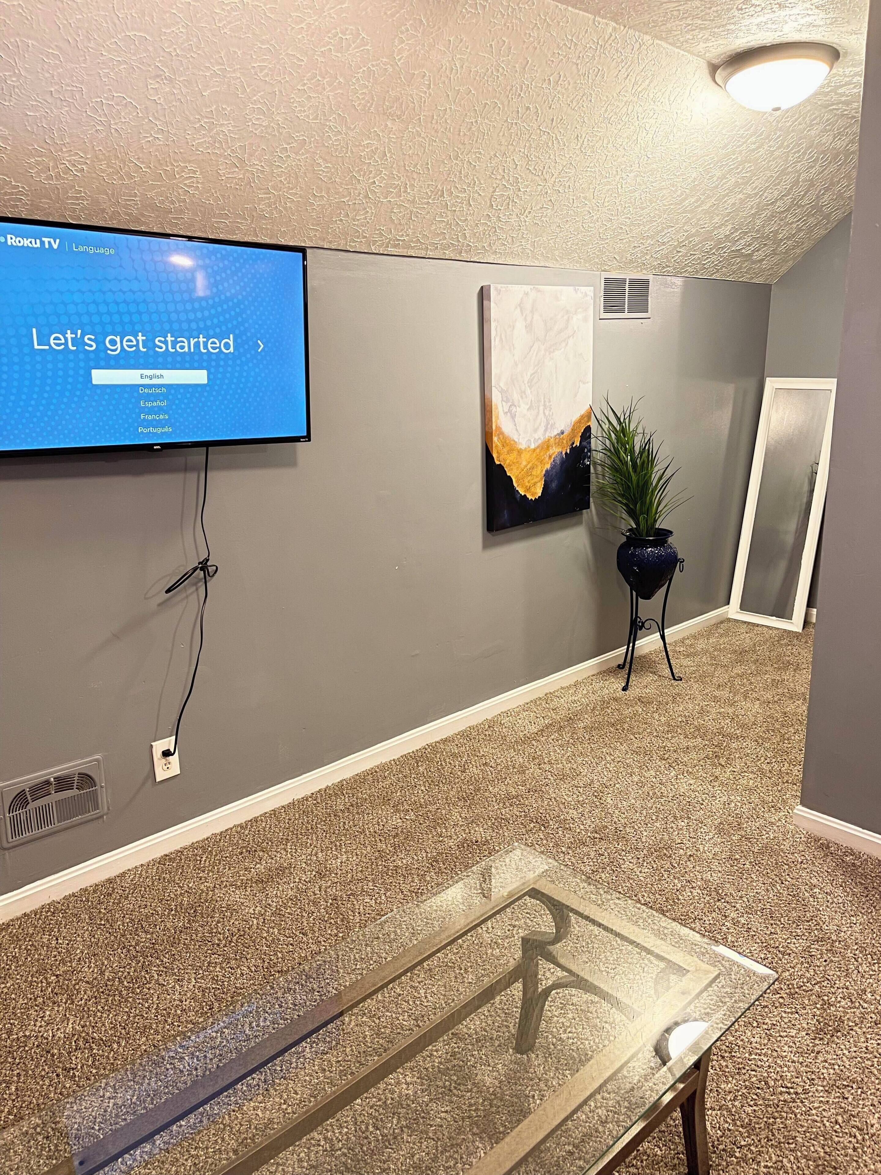 Zona de recepción con un televisor de pantalla plana mostrando un menú de selección de idiomas, una planta en maceta y una obra de arte enmarcada en la pared.
