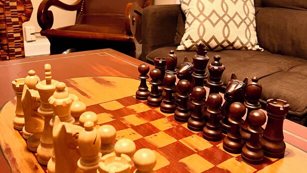 A chessboard set on a wooden table in a living room with a brown leather chair and a grey sofa with patterned cushions.