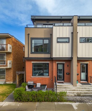 A modern two-story apartment building with a brick facade, large windows, and a small outdoor patio area with a grill. - *Rare find* Calgary’s Finest |Cozy and Vibrant Double King Beds|Stampede Special (Calgary)