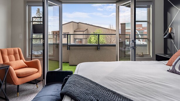 Un dormitorio moderno con una cama grande, una silla naranja y vistas a un edificio a través de puertas correderas abiertas.