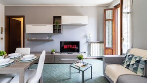 Ein modernes Wohnzimmer mit Essbereich, einem Flachbildfernseher, der 'NETFLIX' anzeigt, und einem Sofa mit einem gemusterten Kissen.