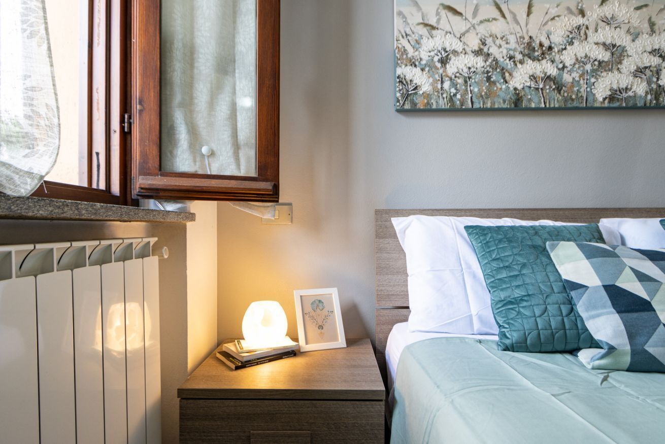 Ein Schlafzimmer mit einem Bett, einem Nachttisch mit Lampe, einem gerahmten Bild und einem Fenster mit Vorhängen.