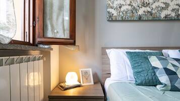 Ein Schlafzimmer mit einem Bett, einem Nachttisch mit Lampe, einem gerahmten Bild und einem Fenster mit Vorhängen.