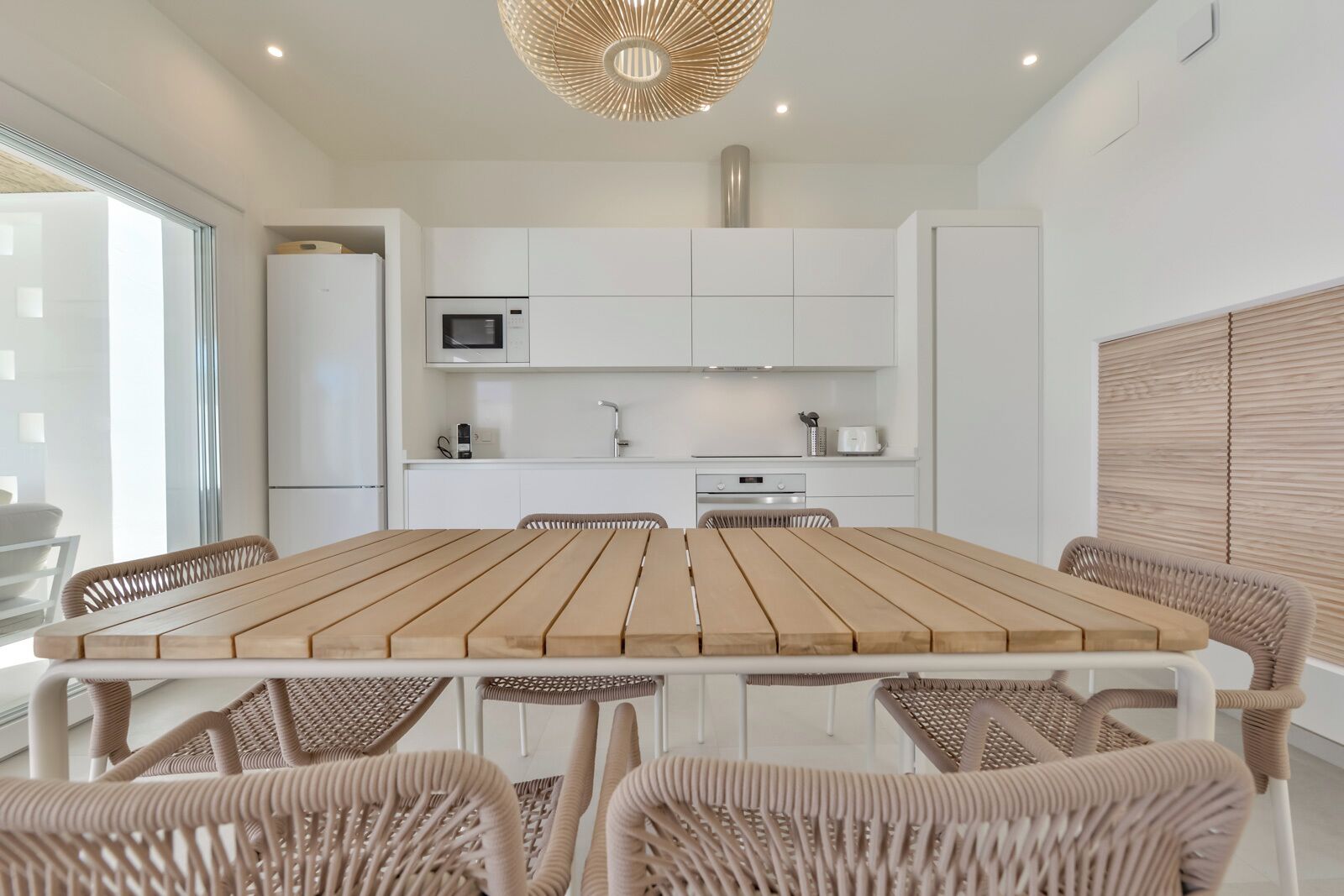 A modern kitchen with white cabinetry, a wooden dining table, wicker chairs, and a unique ceiling light fixture.