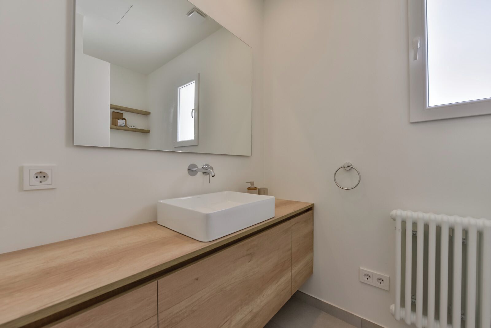 A modern bathroom with a white sink, wooden vanity, a large mirror, and a radiator.