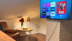 A living room with a flat-screen TV mounted on a brick wall, a brown leather couch, a small round table with a lamp, and a vase with red flowers. - Near Airport & Major Attractions, 5Mi Downtown, Comfy Loft (Louisville)