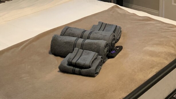 A neatly made bed with rolled towels, a small desk with a lamp, a chair, and a window with blinds.
