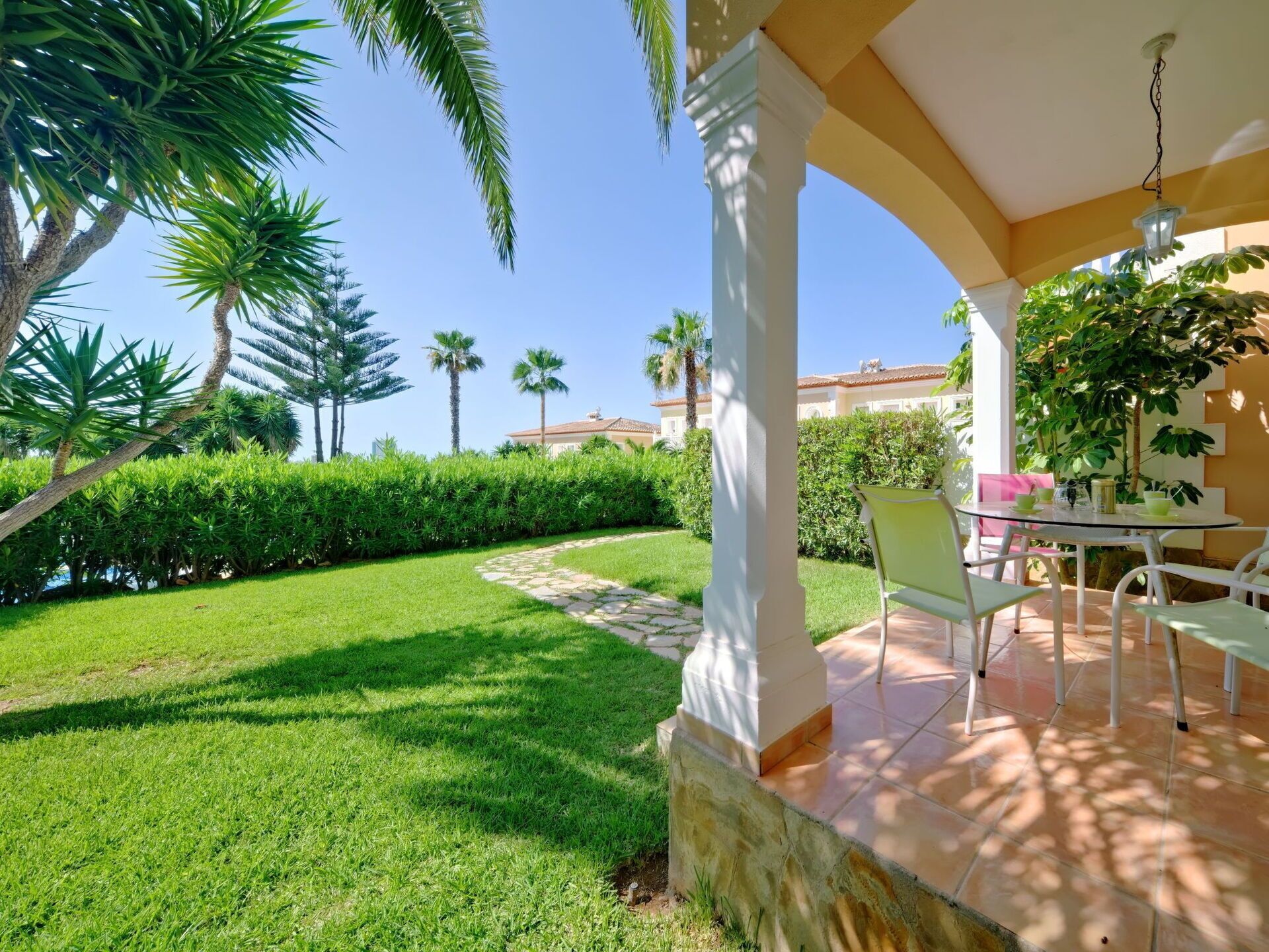 A covered outdoor area with a patio table and chairs, surrounded by lush greenery and palm trees.