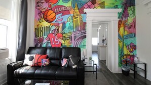 Una sala de estar con un sofá de cuero negro, arte mural colorido y una cocina moderna al fondo.