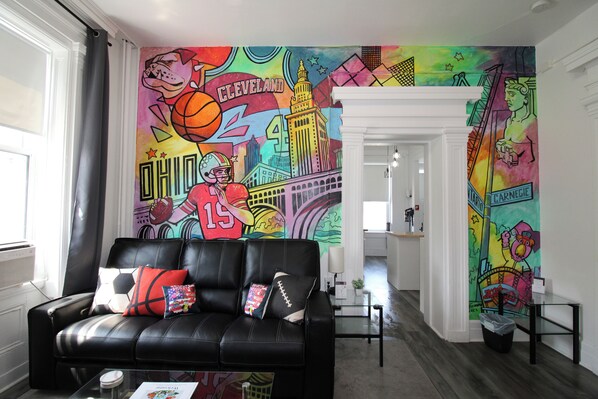A living room with a black leather sofa, colorful wall art, and a modern kitchen in the background.