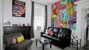 Una sala de estar con un sofá de cuero negro, una mesa de centro de vidrio, una chimenea y un mural de colores en la pared.
