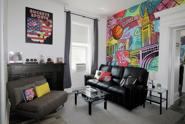 Una sala de estar con un sofá de cuero negro, una mesa de centro de vidrio, una chimenea y un mural de colores en la pared.