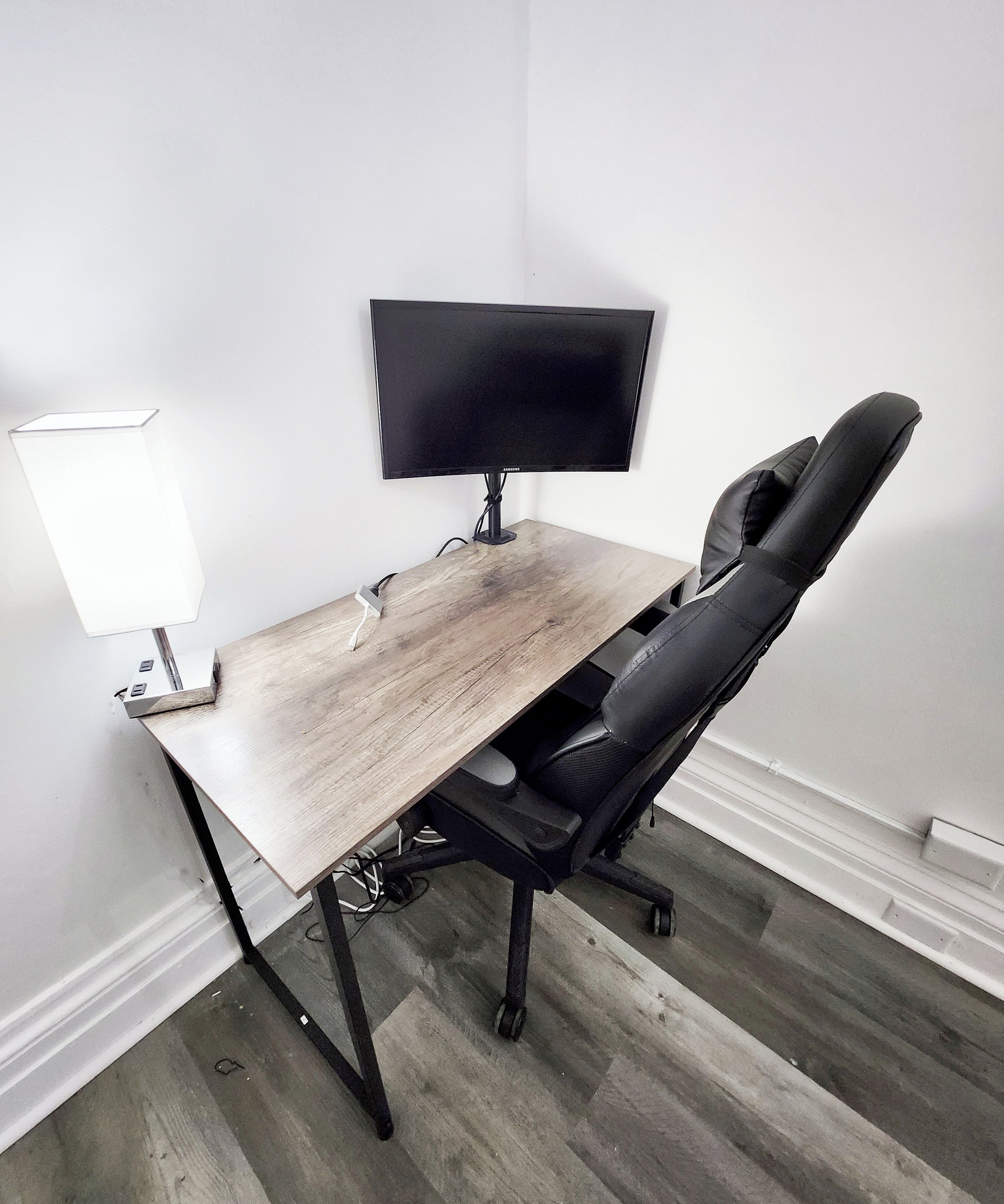 Un bureau d’angle avec un bureau en bois, un fauteuil de bureau noir et un écran plat fixé au mur.