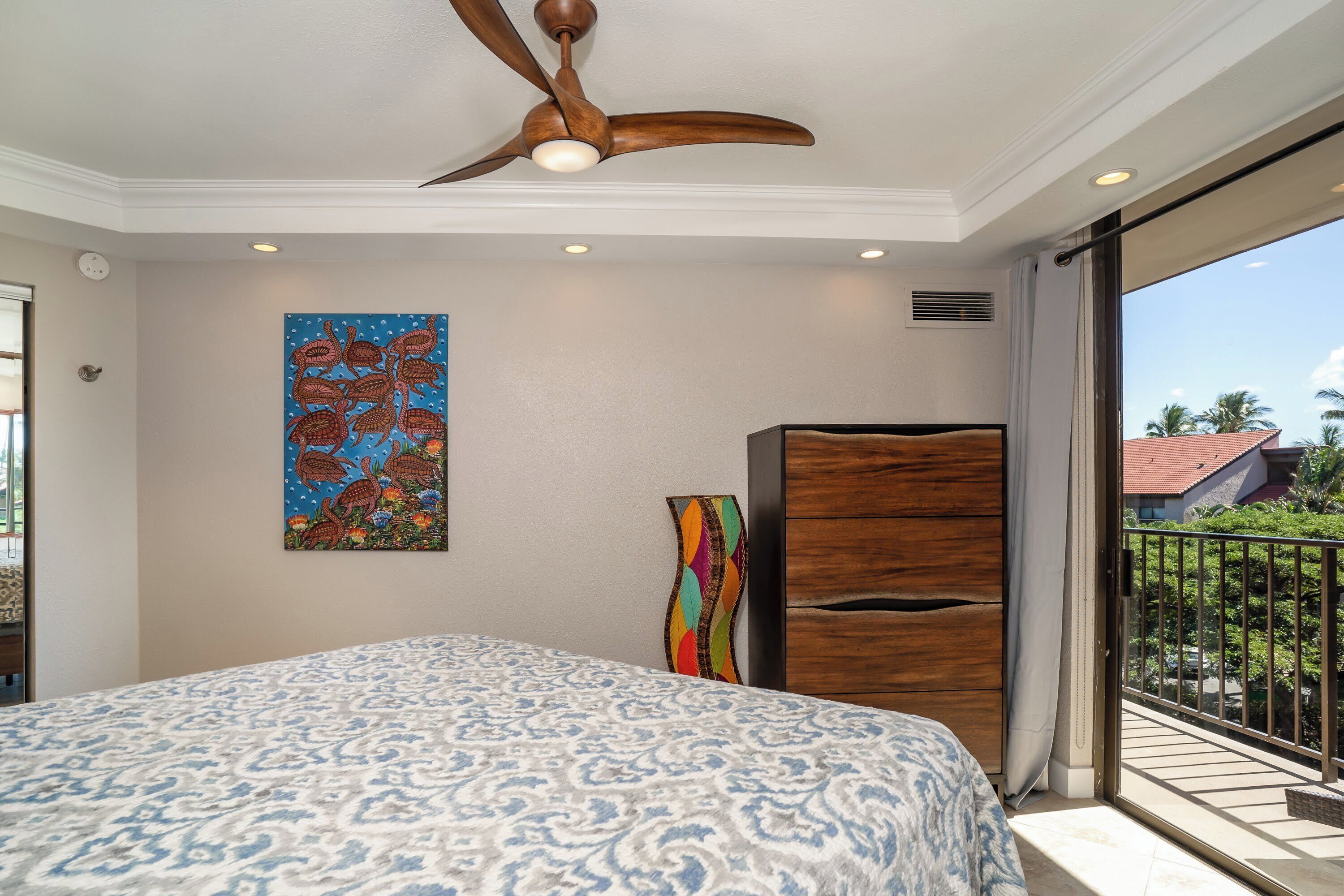 Ein Schlafzimmer mit einem großen Bett, einem Deckenventilator, einem Gemälde, einer Holzkabine und einem Balkon mit Blick auf Palmen.