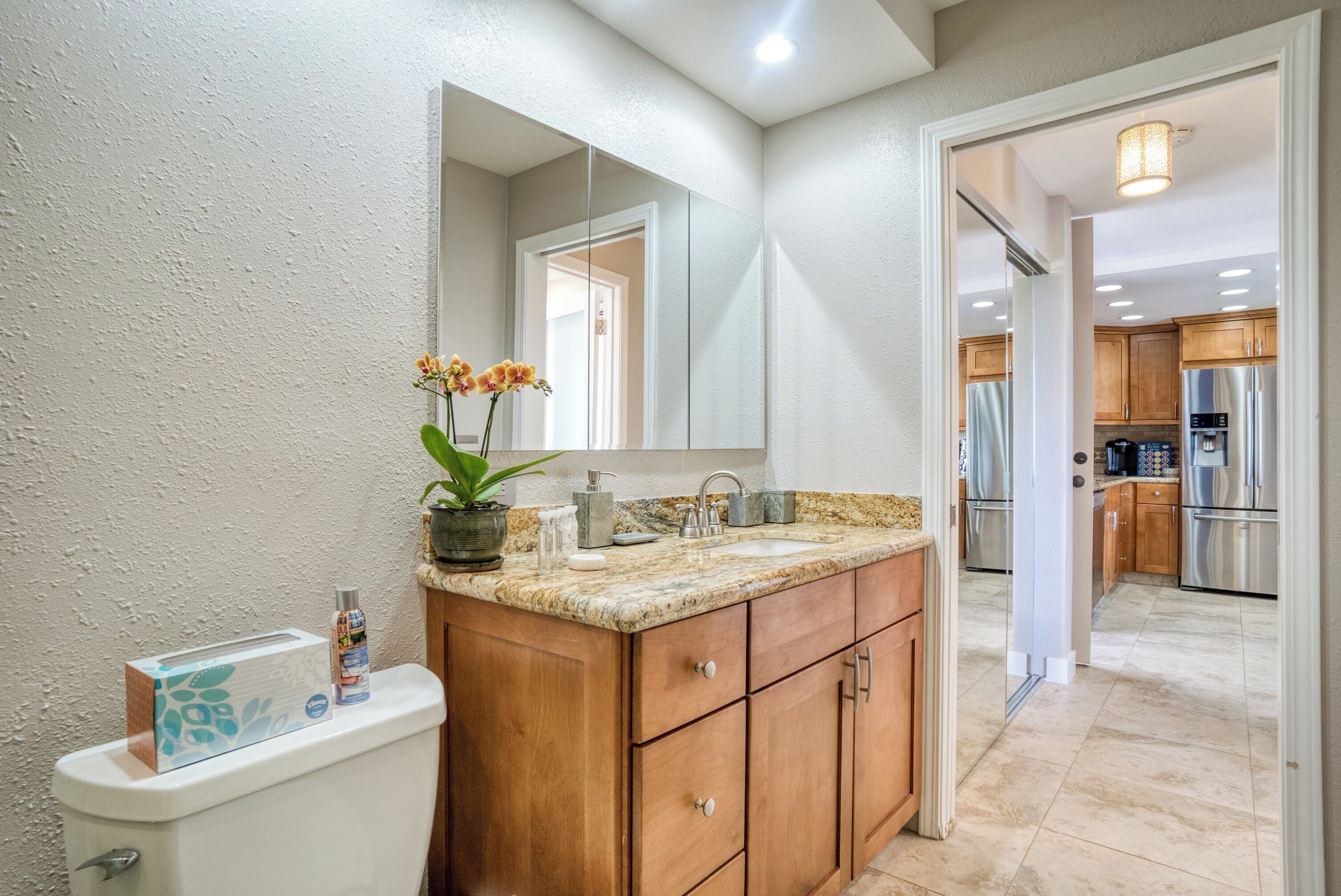 Une salle de bain avec un meuble-lavabo en bois, un évier et un miroir. On trouve des toilettes sur la gauche et une porte qui donne sur la cuisine.