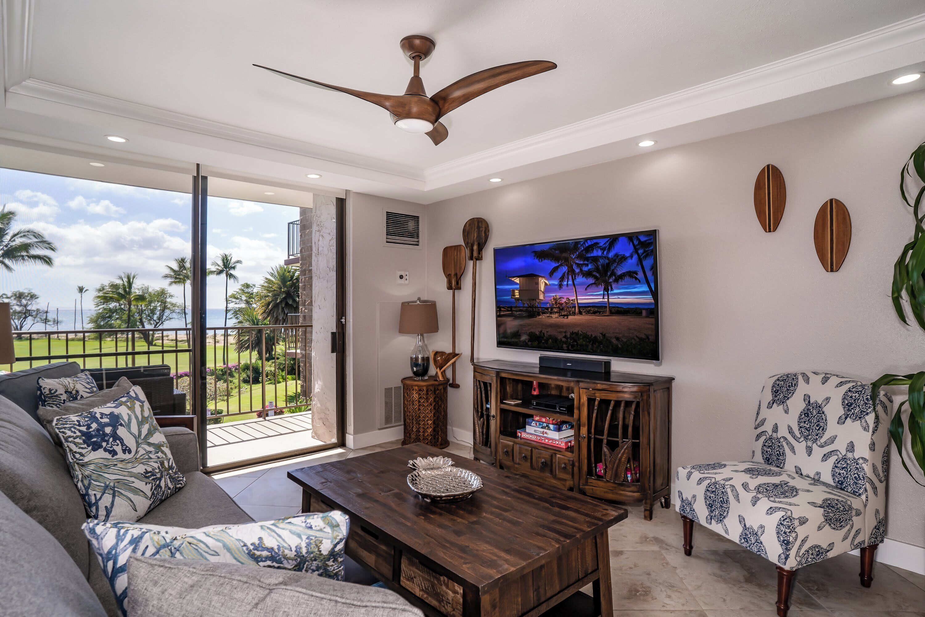 A living room with a sofa, armchair, coffee table, TV, and a balcony with a view of palm trees and a golf course.