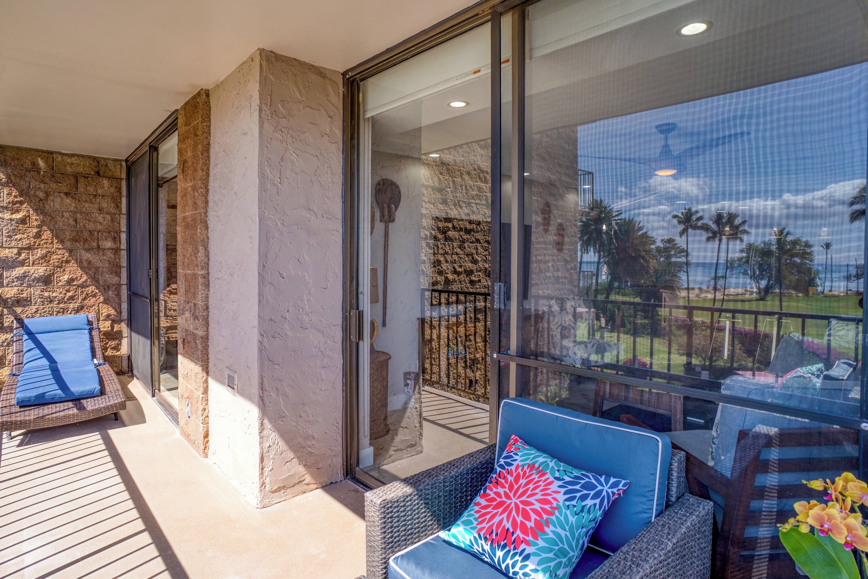 A balcony with wicker furniture, a glass door leading to a pool area, and a view of palm trees and a clear sky.