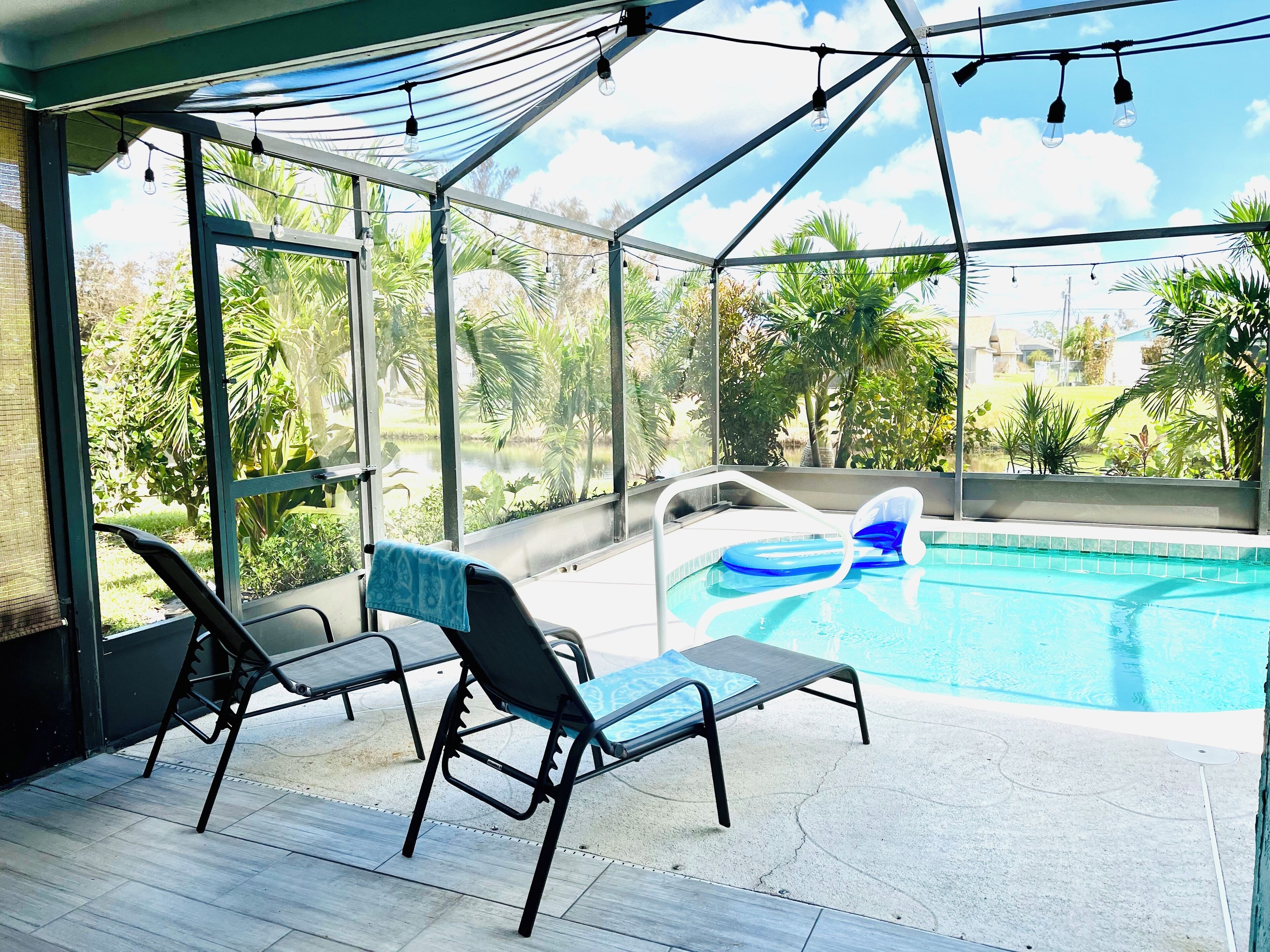 A covered outdoor area with a pool, lounge chairs, and a view of greenery.