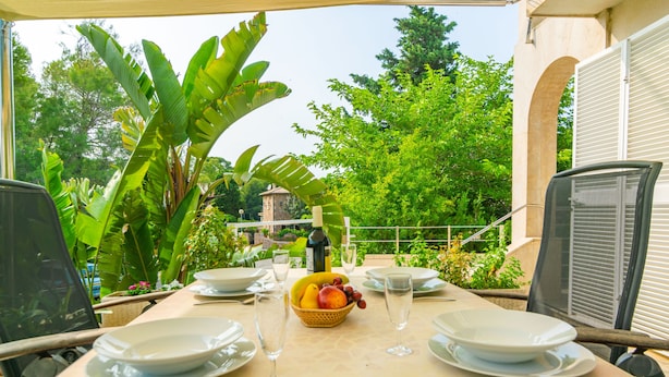 Un área de comedor al aire libre con una mesa preparada para dos personas, rodeada de exuberante vegetación y vista a un edificio al fondo.