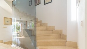 Una escalera con peldaños color beige y barandilla de vidrio, que conduce a una habitación con cama y ventana.
