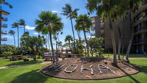 A landscaped area with a circular stone design spelling "MOHAI" surrounded by palm trees and a building with balconies. - Modern and Relaxing Oceanfront Vistas (Kihei)