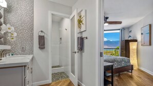 A modern bedroom with a wooden floor, a bed with a patterned bedspread, a bathroom with a tiled shower, and a balcony with a view of the mountains. - Modern and Relaxing Oceanfront Vistas (Kihei)