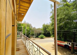 Une terrasse en bois avec une rambarde en métal, donnant sur un chemin de terre et un véhicule rouge.