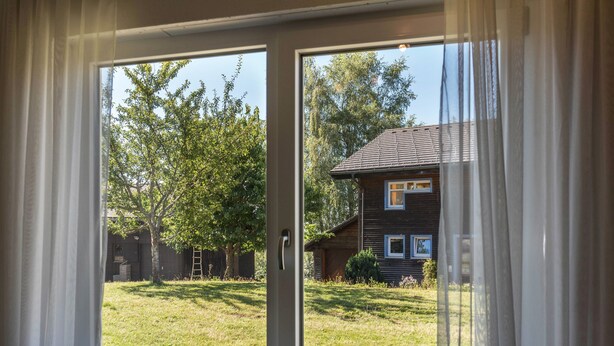 A view through a window with curtains, revealing a house, a grassy yard, and trees. - Apartment 'Haus Isele' with Mountain View, Terrace and Wi-Fi (Dachsberg)