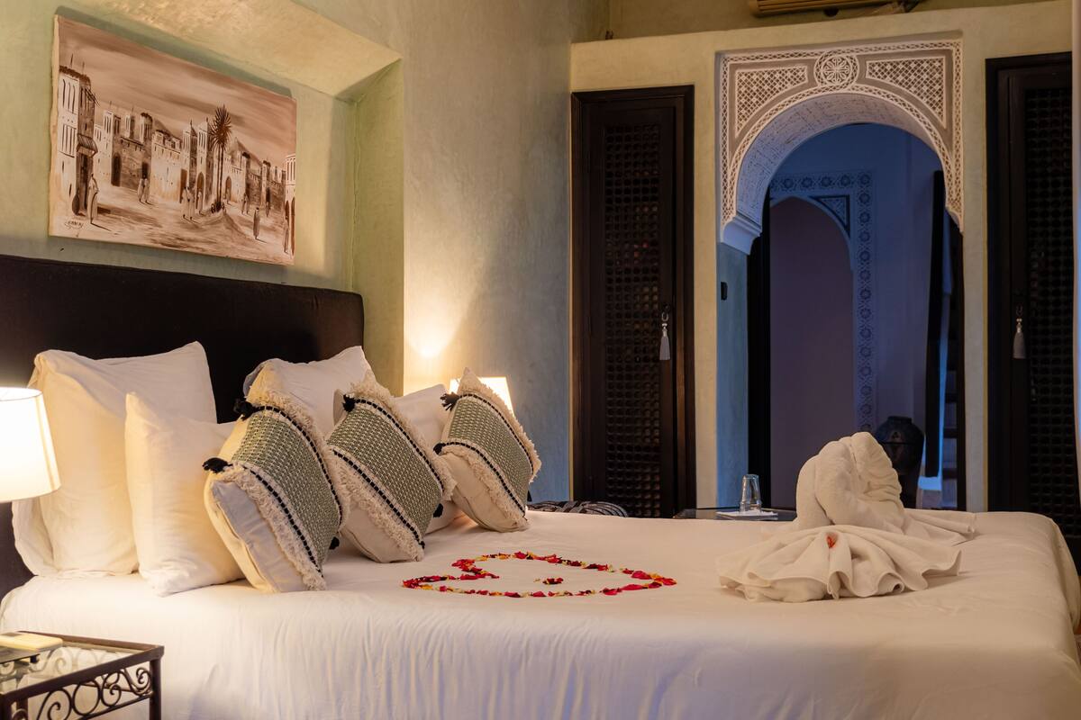 Une chambre d’hôtel avec un grand lit, une tête de lit, une table de chevet, une lampe, un tableau au mur et une porte en arc.