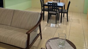 Ein Wohnzimmer mit einer Couch, einem Esstisch mit Stühlen, einer Küchenzeile mit Holzschränken und einem Fenster mit Vorhängen.