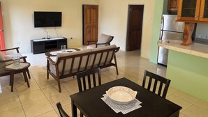 Ein Wohnzimmer mit einer Couch, einem Esstisch mit einer Schüssel, einem Fernseher und einer Küchenzeile mit einem Kühlschrank.