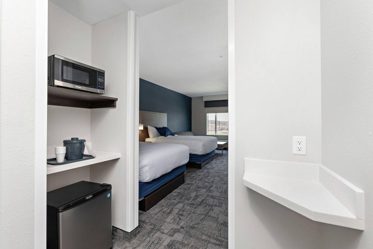 A hotel room with two beds, a microwave, a mini fridge, and a white shelf.
