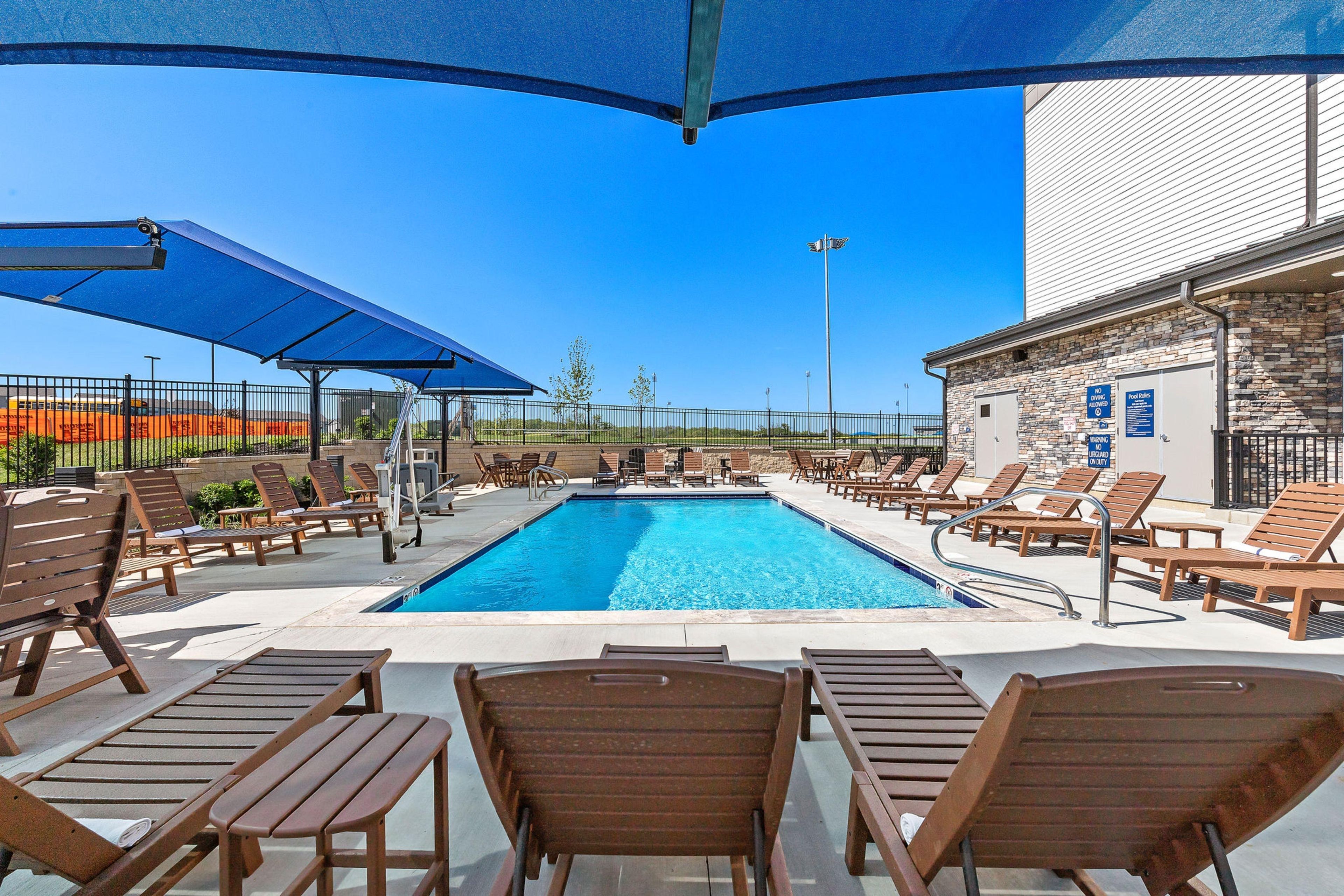 Un espace piscine avec des chaises longues et des parasols, un bâtiment en pierre et, au fond, un terrain de sport délimité par une clôture.