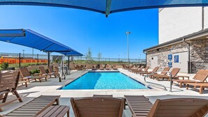 Ein Poolbereich mit Liegestühlen und Sonnenschirmen, ein Steinhaus und im Hintergrund ein eingezäunter Sportplatz.
