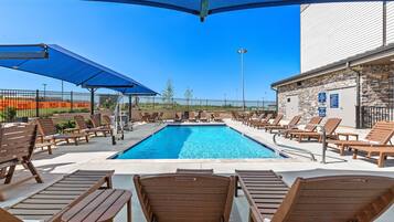 Un espace piscine avec des chaises longues et des parasols, un bâtiment en pierre et, au fond, un terrain de sport délimité par une clôture.