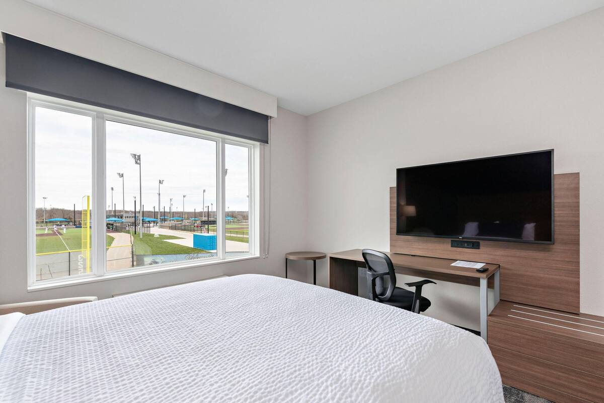 A hotel room with a large bed, a desk with a chair, a flat-screen TV mounted on the wall, and a window offering a view of a sports field.