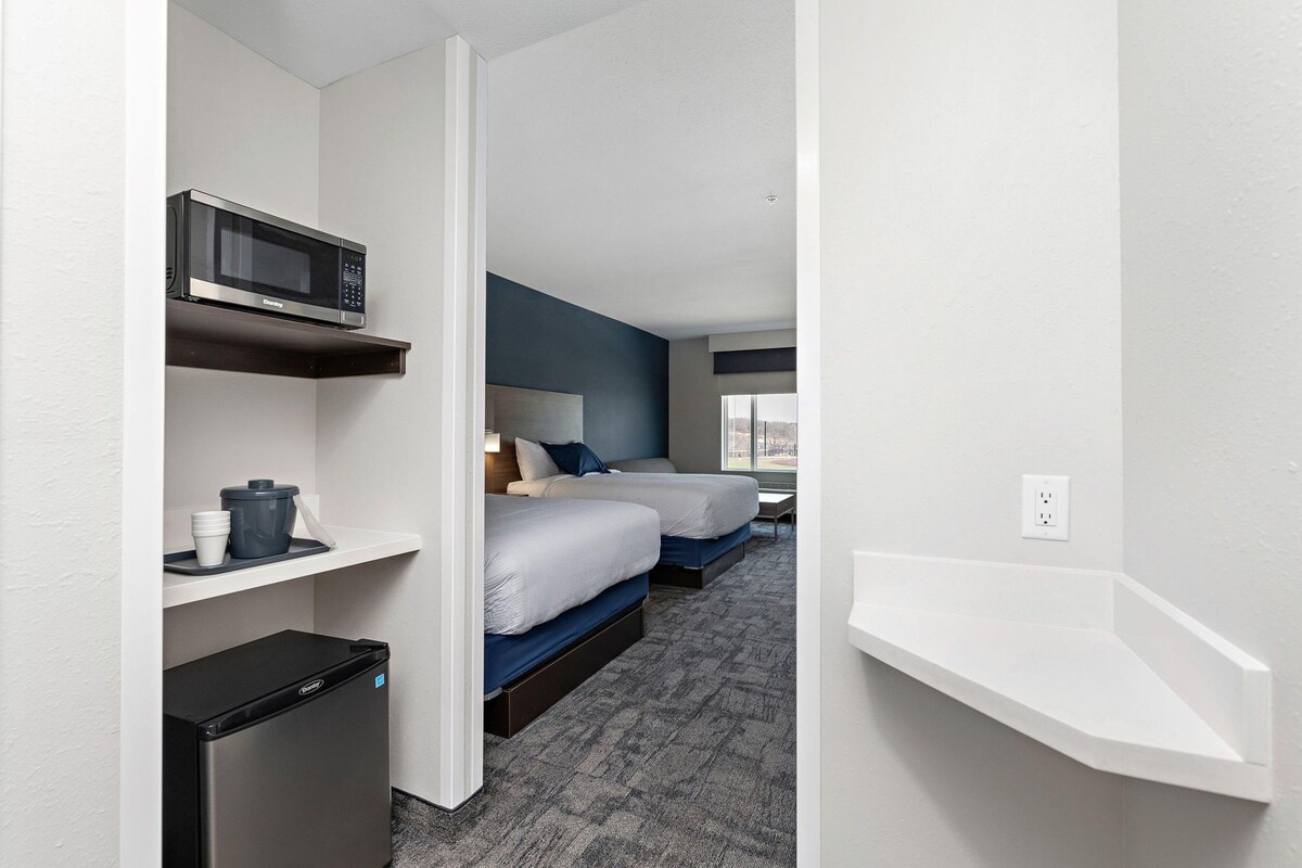 A hotel room with two beds, a microwave, a mini fridge, and a white shelf.