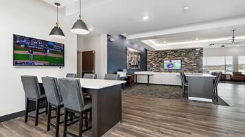 A modern, well-lit room with a large flat-screen TV displaying a baseball game, a kitchen area with a stone backsplash, and a seating area with a dark wood floor.