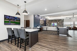 A modern, well-lit room with a large flat-screen TV displaying a baseball game, a kitchen area with a stone backsplash, and a seating area with a dark wood floor. - Holiday Inn Express Kansas City North Parkville (Parkville)
