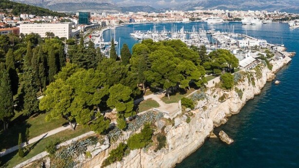 An aerial view of a coastal area with a marina, rocky cliffs, and dense greenery. - Beachfront apartment with balcony and parking (Split)