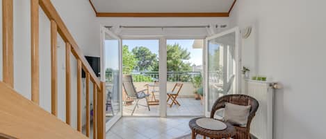 A room with a wooden staircase, a wicker chair, a small table, and a view of an outdoor balcony with chairs and plants.