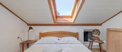A bedroom with a skylight, a bed with white linens, a bedside lamp, a wooden chair, and a wardrobe.